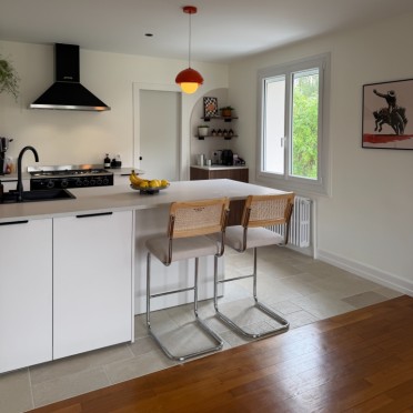 Cuisine ouverte moderne avec îlot central blanc, suspension ronde orange, et deux chaises de bar en cannage sur pieds chromés