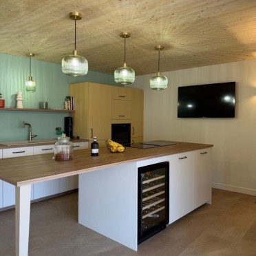 Cuisine ouverte avec îlot central bois et blanc, suspensions vertes en laiton, façades ocre et crédence vert d'eau.